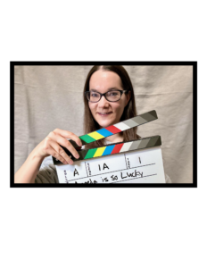 Photograph An image of Dr. Kim Maddison standing in front of a beige backdrop, holding a clapper board marked 'Roll A, Scene 1A, Take 1, Angela is so Lucky'.