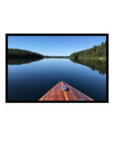 Photograph, colour The wooden bow of a boat, showing some age, heading toward a passage of perfectly still water lined on both sides with banks of coniferous trees, beneath a rich blue sky. The sky and treelines are mirrored in the water.