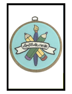 Embroidery in Loop on white backdrop. A blue cloth, with embroidered image of two crossed pencils, one yellow, one green, and a blue paintbrush behind both, with a white ribbon reading 'don't hate, create' on it.