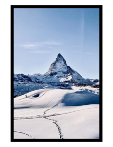 Photograph: a solitary mountain in the distance against a pale blue sky. The foreground is covered in snow, with rolling hills and lower peaks in the middle distance. A solitary hiker silhouetted against the white snow walks toward the mountain.