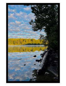 Photograph. A bright morning with a lake shore silhouetted in right foreground. The lake is perfectly still, and reflects green coniferous trees on the far bank, as well as the sky, dotted with clouds.